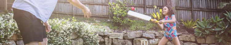 young girl hitting cricket ball thrown by man in yard
