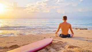 Man meditating on beach next to surfboard