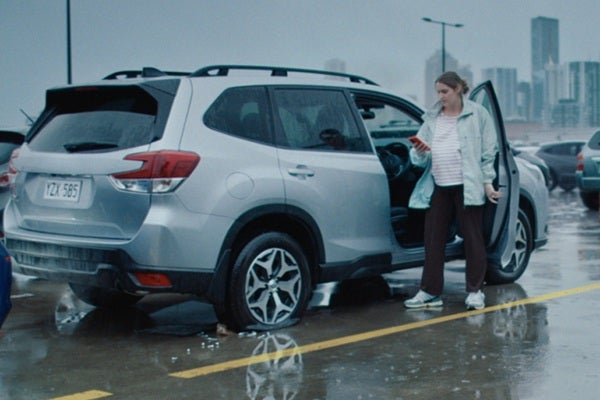 In a carpark, a woman is standing in the rain beside her car which has a flat tyre. She is looking at her phone.