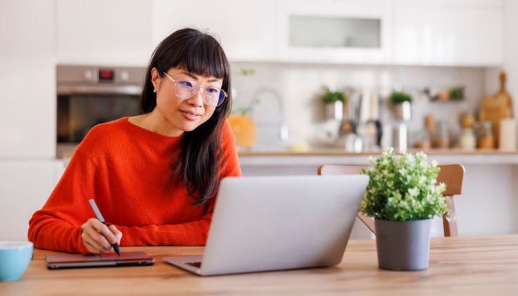 Woman sitting at kitchen table looking at laptop while writing on a tablet.