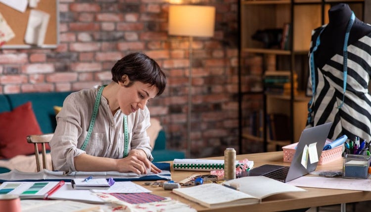 Fashion designer with short brown hair is sitting at a table surrounded by notes, swatches and sewing equipment.