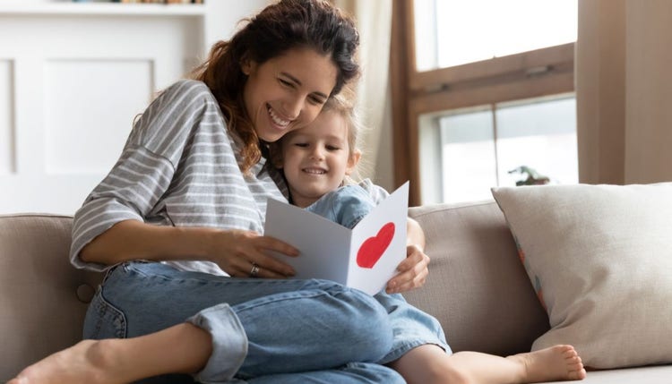 A woman and young girl are snuggled together on a couch. They are both smiling as they read a card with a red love heart on the front/