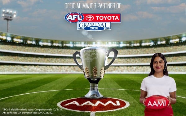 The AFL premiership cup sits on the ground of the MCG. A woman with dark hair and a white t-shirt stands in the foreground holding a football that says AAMI