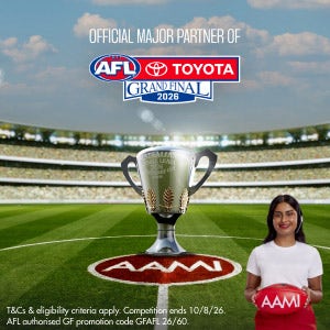 The AFL premiership cup sit on the ground of the MCG. A woman with dark hair wearing a white t-shirt stands in the foreground holding a football that says AAMI